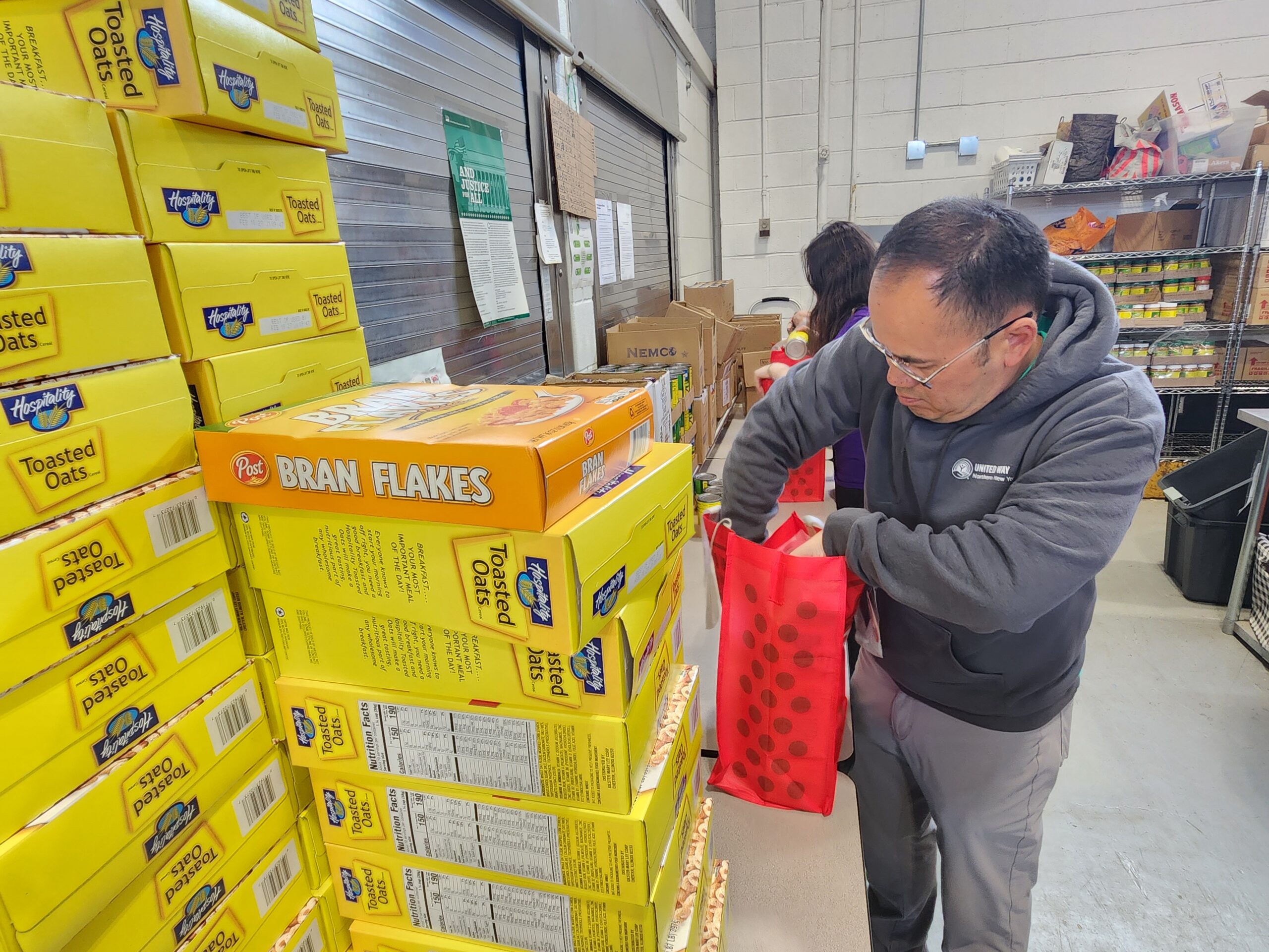 Large stack of cereal boxes in foreground, man packing food bag.