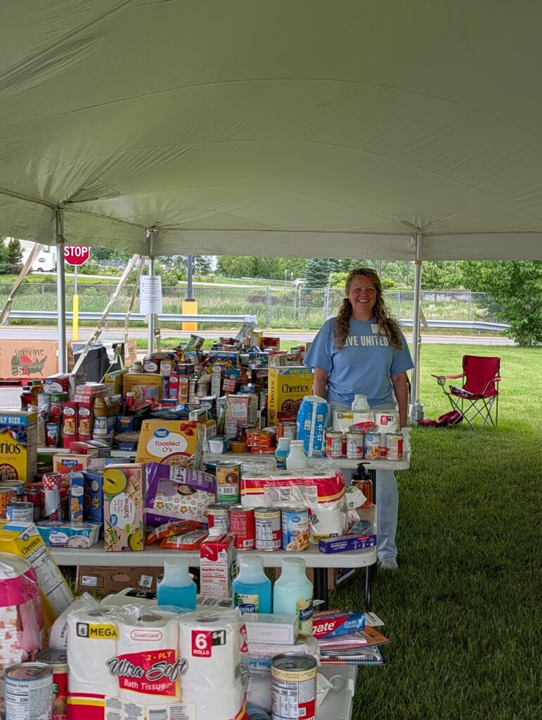 Volunteer with blue LIVE UNITED shirt stands inside tent with tables full of food.