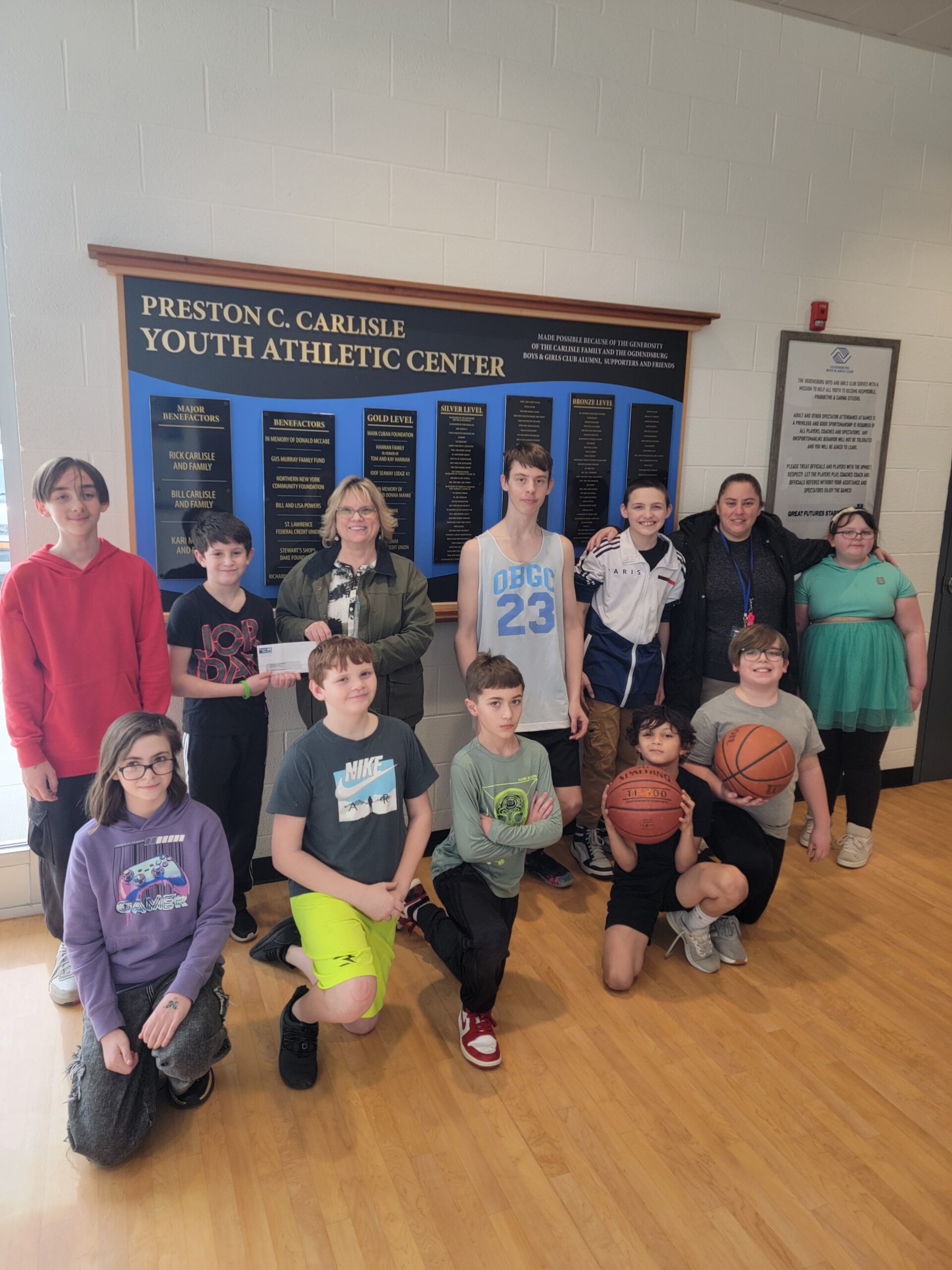 Youth at the Ogdensburg Boys and Girls Club strike a pose and accept a check from UWNNY CEO Dawn Cole. A sign in the background promotes the Preston C. Carlisle Youth Athletic Center.