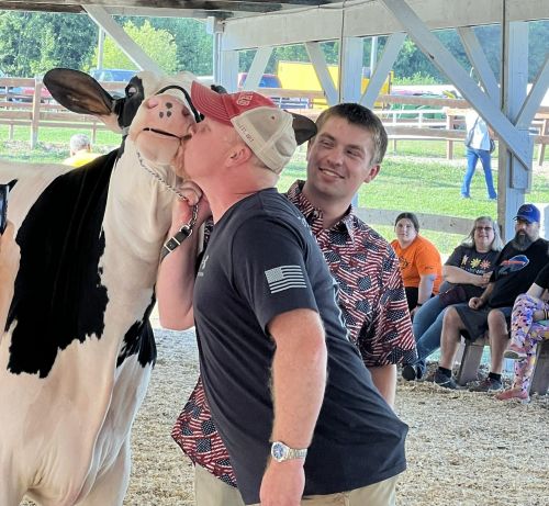 2026 Cow Kissing winner Mike Jackson kisses a cow while the handler looks on and smiles.